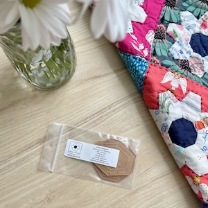 May include: A clear plastic bag containing a brown plastic template for quilting. The template is labelled "Casey Large 629" and has a 3/8" seam allowance. The bag is on a wooden surface with a colourful quilt in the background.