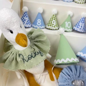 May include: A plush white goose toy with a yellow beak, wearing a sage green ruffled collar embroidered with the name Rocky. The set includes a tiered display stand holding twelve small numbered party hats and a matching blue ruffled collar.