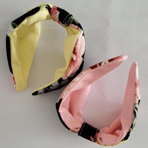 May include: Two fabric headbands. One is black with a floral pattern and a yellow interior. The other is pink with a floral pattern and a pink interior. Both headbands have a knotted design.