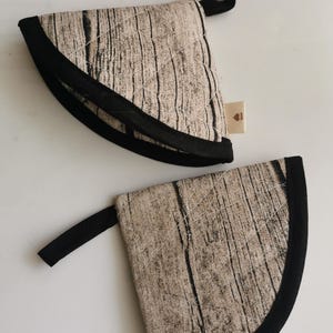 Könnte beinhalten: Zwei braune und schwarze gepolsterte Topflappen mit Holzmaserung. Die Topflappen haben einen schwarzen Rand und eine Aufhängeschlaufe.