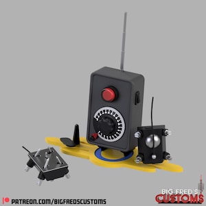 May include: A black and gray remote control device with a red button, dial, and antenna. The device is mounted on a yellow base with a blue ring. Additional components are also present. The text "BIG FRED'S CUSTOMS" and a website address are visible.