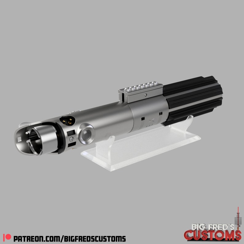 Pode incluir: Um adere&ccedil;o de sabre de luz prateado com uma al&ccedil;a preta e um suporte de pl&aacute;stico transparente. O sabre de luz tem um corpo de metal prateado com uma al&ccedil;a preta e uma ponta de metal prateado. O suporte &eacute; de pl&aacute;stico transparente e tem uma base retangular.