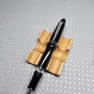 Puede incluir: Una pluma estilogr&aacute;fica negra con detalles plateados y un plum&iacute;n plateado descansa sobre un soporte de madera para bol&iacute;grafos. La pluma tiene un acabado brillante y una secci&oacute;n transparente cerca del plum&iacute;n. El soporte de madera muestra la veta natural.