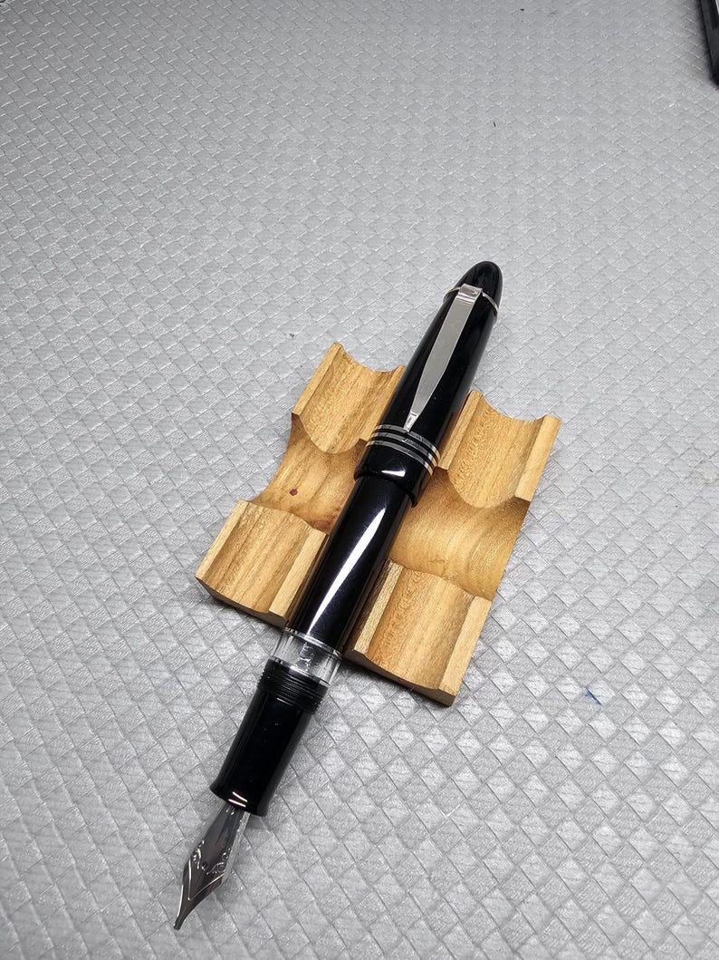 Puede incluir: Una pluma estilogr&aacute;fica negra con detalles plateados y un plum&iacute;n plateado descansa sobre un soporte de madera para bol&iacute;grafos. La pluma tiene un acabado brillante y una secci&oacute;n transparente cerca del plum&iacute;n. El soporte de madera muestra la veta natural.