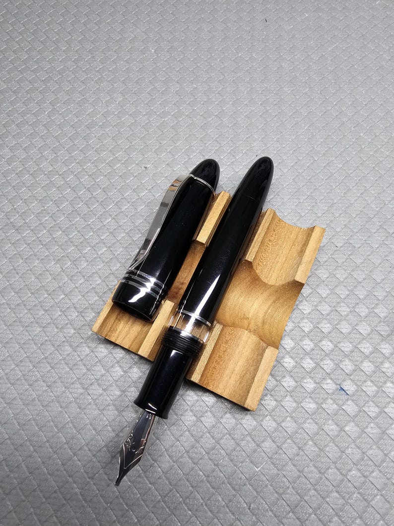 Puede incluir: Una pluma estilogr&aacute;fica negra con detalles plateados y un plum&iacute;n plateado descansa sobre un soporte de madera para bol&iacute;grafos. La tapa de la pluma est&aacute; abierta, revelando el plum&iacute;n. El soporte para bol&iacute;grafos es de madera clara.