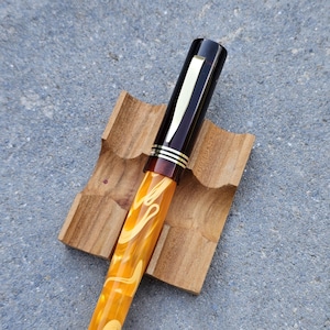 Könnte beinhalten: Ein Füllfederhalter mit einem marmorierten gelb-orangen Harzschaft und einer dunkelbraunen Kappe. Die Kappe verfügt über einen goldfarbenen Metallclip und drei dünne goldfarbene Ringe an der Basis. Der Stift liegt in einem kleinen, quadratischen Holzständer.