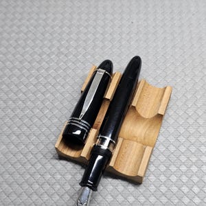 Puede incluir: Una pluma estilogr&aacute;fica negra con detalles plateados, colocada sobre un soporte de madera. La tapa de la pluma est&aacute; quitada, revelando el plum&iacute;n. El soporte es de madera clara.