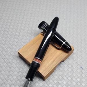 Puede incluir: Una pluma estilogr&aacute;fica negra con detalles plateados y una banda naranja, apoyada sobre un peque&ntilde;o bloque de madera. Se ve el plum&iacute;n y el capuch&oacute;n est&aacute; quitado. Un instrumento de escritura cl&aacute;sico, s&iacute;mbolo de elegancia.