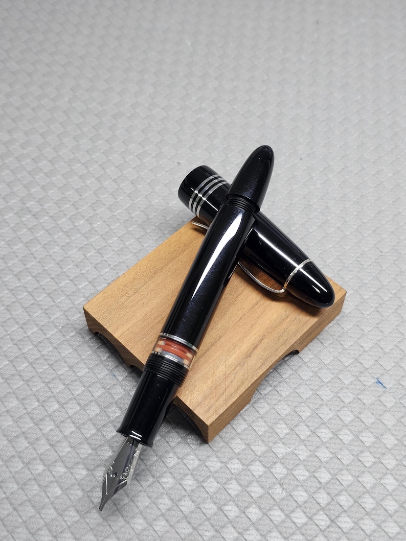 Puede incluir: Una pluma estilogr&aacute;fica negra con detalles plateados y una banda naranja, apoyada sobre un peque&ntilde;o bloque de madera. Se ve el plum&iacute;n y el capuch&oacute;n est&aacute; quitado. Un instrumento de escritura cl&aacute;sico, s&iacute;mbolo de elegancia.