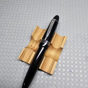 Puede incluir: Una pluma estilogr&aacute;fica negra con detalles plateados descansa sobre un peque&ntilde;o soporte de madera. La pluma tiene un acabado brillante y un clip plateado. El soporte para la pluma es de madera clara con veta natural.