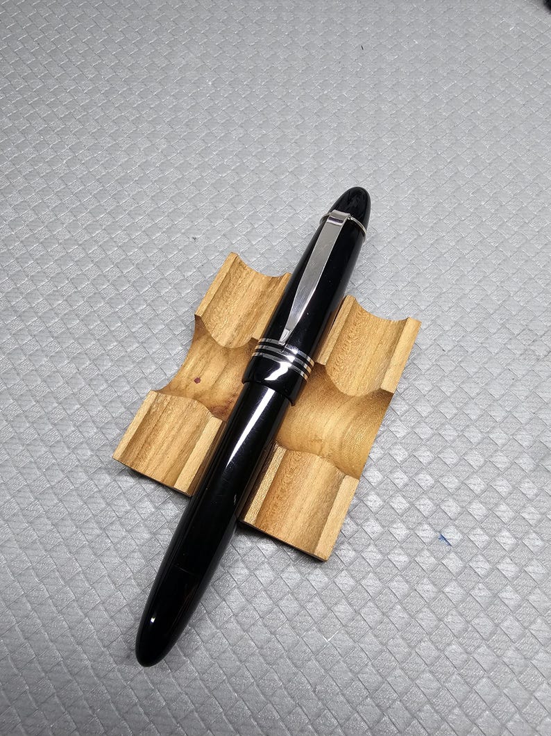 Puede incluir: Una pluma estilogr&aacute;fica negra con detalles plateados descansa sobre un peque&ntilde;o soporte de madera. La pluma tiene un acabado brillante y un clip plateado. El soporte para la pluma es de madera clara con veta natural.