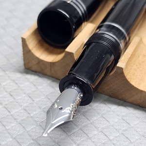 Puede incluir: Una pluma estilogr&aacute;fica negra con detalles plateados, colocada en un soporte de madera. La tapa est&aacute; quitada, mostrando la c&aacute;mara de tinta. El plum&iacute;n tiene grabados decorativos.