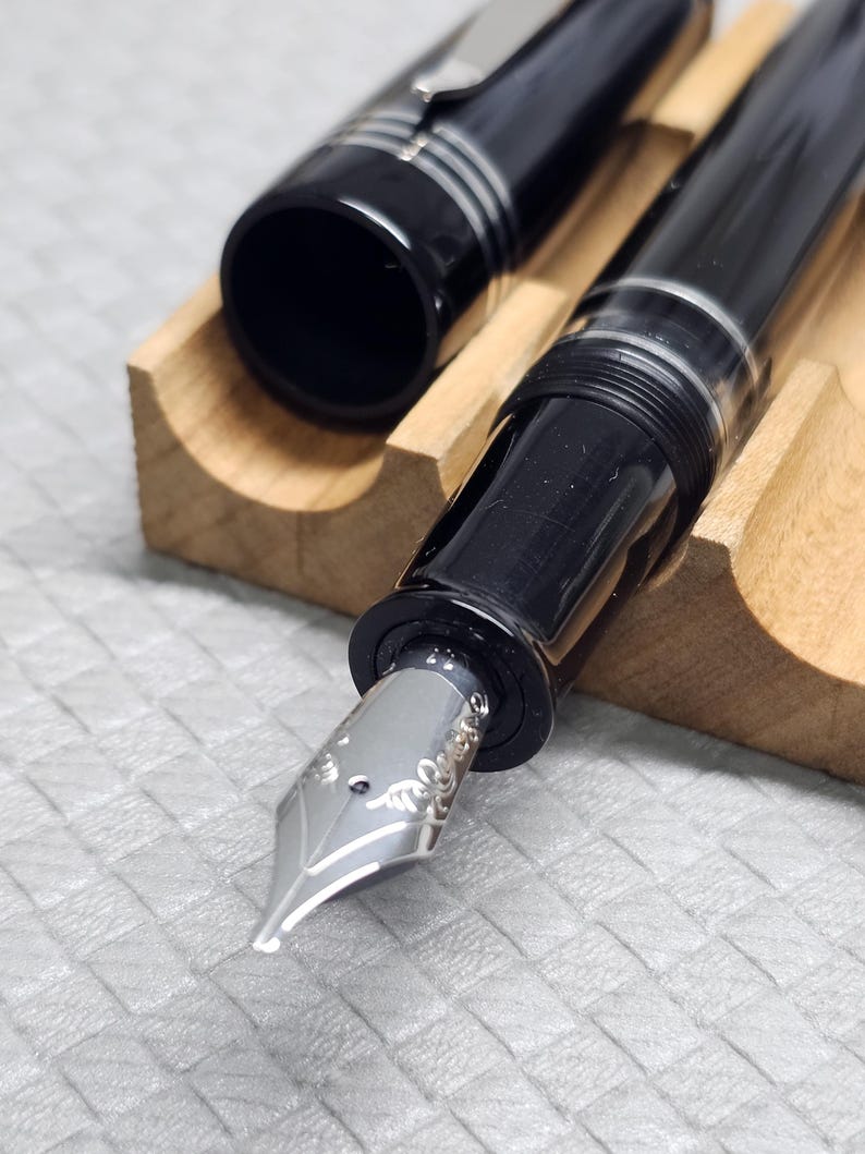 Puede incluir: Una pluma estilogr&aacute;fica negra con detalles plateados, colocada en un soporte de madera. La tapa est&aacute; quitada, mostrando la c&aacute;mara de tinta. El plum&iacute;n tiene grabados decorativos.