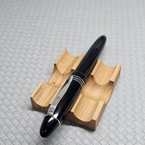 Puede incluir: Una pluma estilogr&aacute;fica negra con detalles plateados y un clip plateado descansa sobre un soporte de madera para bol&iacute;grafos. El bol&iacute;grafo tiene un acabado brillante y un dise&ntilde;o aerodin&aacute;mico. El soporte para bol&iacute;grafos es de madera clara.