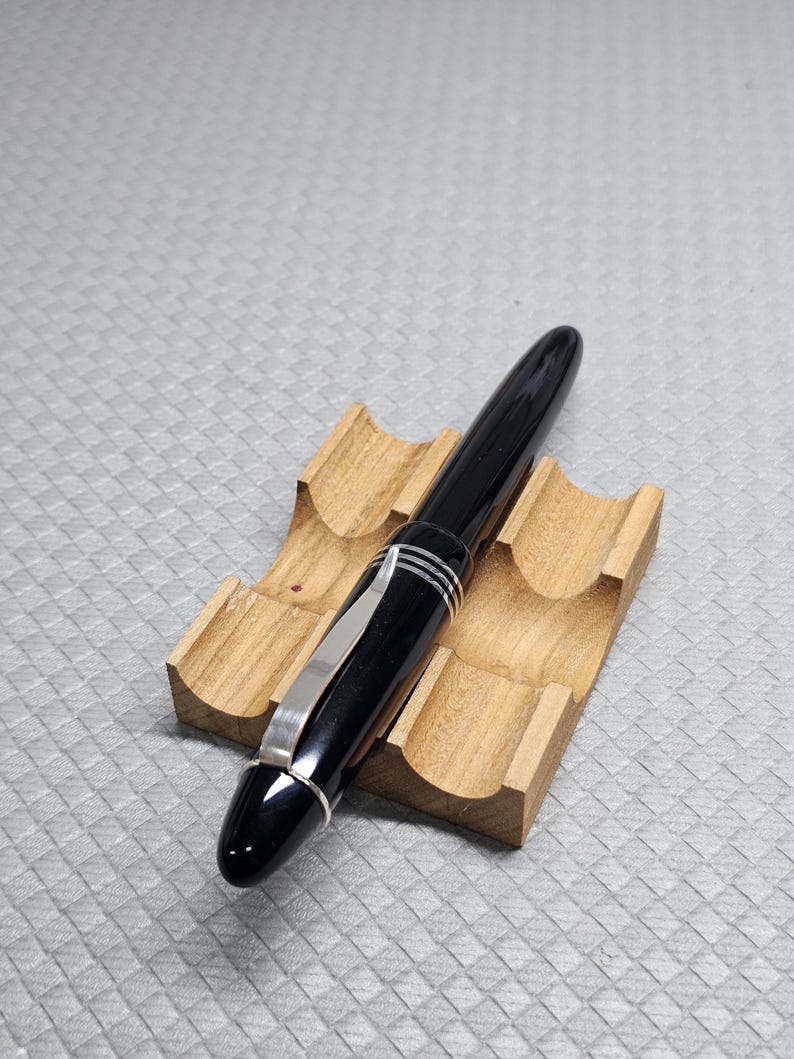 Puede incluir: Una pluma estilogr&aacute;fica negra con detalles plateados y un clip plateado descansa sobre un soporte de madera para bol&iacute;grafos. El bol&iacute;grafo tiene un acabado brillante y un dise&ntilde;o aerodin&aacute;mico. El soporte para bol&iacute;grafos es de madera clara.