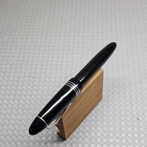 Puede incluir: Una elegante pluma estilogr&aacute;fica negra con detalles plateados y un clip plateado, presentada sobre un peque&ntilde;o soporte de madera. La pluma tiene tres bandas plateadas y un dise&ntilde;o aerodin&aacute;mico. Fondo gris texturizado.