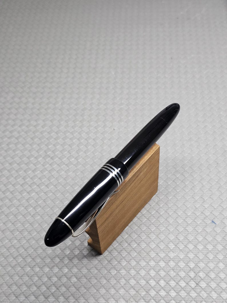 Puede incluir: Una elegante pluma estilogr&aacute;fica negra con detalles plateados y un clip plateado, presentada sobre un peque&ntilde;o soporte de madera. La pluma tiene tres bandas plateadas y un dise&ntilde;o aerodin&aacute;mico. Fondo gris texturizado.