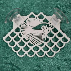 Op de afbeelding: Witte plastic aquariumdecoratie met zuignappen. De decoratie heeft een gestileerde vis met schubben en een decoratieve rand.