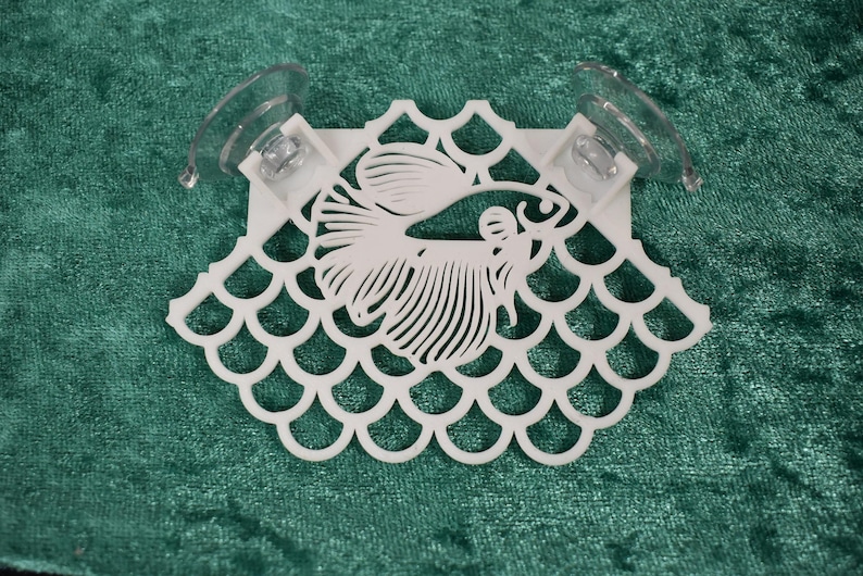 Op de afbeelding: Witte plastic aquariumdecoratie met zuignappen. De decoratie heeft een gestileerde vis met schubben en een decoratieve rand.