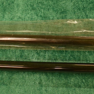 Könnte beinhalten: Zwei dunkelbraune zylindrische Röhren, eine in einer transparenten Kunststoffhülle, und ein Produktetikett mit dem Markennamen "Fluval" und Modellnummern. Die Röhren sind wahrscheinlich Aquarienfilterteile, die auf einer grünen Oberfläche liegen.