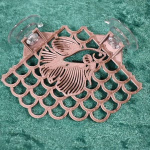 Op de afbeelding: Een roze houten vis-uitsparing met een decoratief schubbenpatroon, bevestigd aan twee transparante zuignappen.