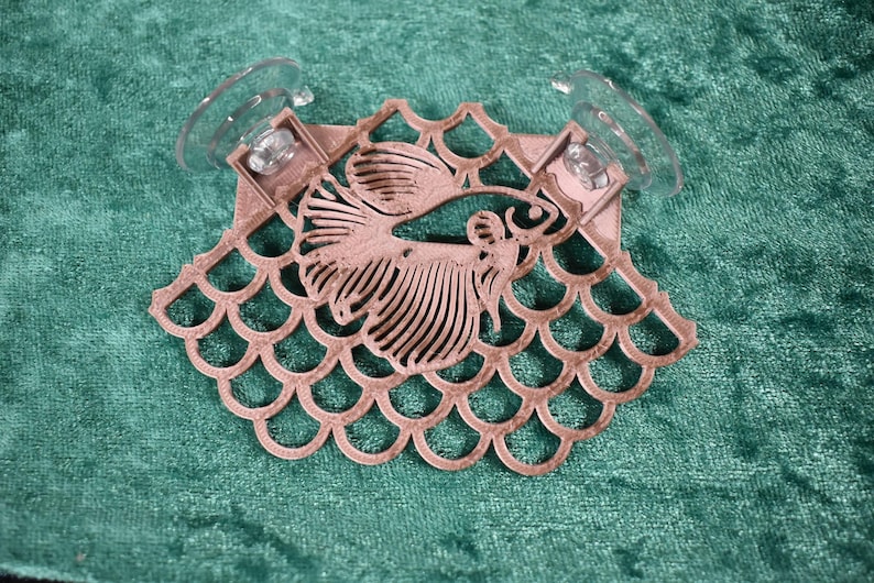 Op de afbeelding: Een roze houten vis-uitsparing met een decoratief schubbenpatroon, bevestigd aan twee transparante zuignappen.