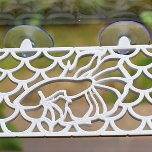 Op de afbeelding: Witte plastic aquariumdecoratie met een zuignap aan elke kant. De decoratie toont een vis met een schubbenpatroon.