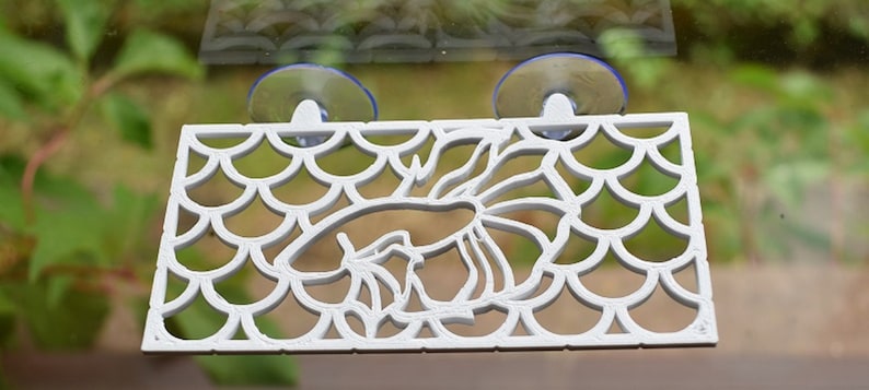 Op de afbeelding: Witte plastic aquariumdecoratie met een zuignap aan elke kant. De decoratie toont een vis met een schubbenpatroon.