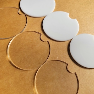 May include: Four white acrylic coasters with a cut-out for a cup. The coasters are on a brown surface.