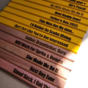 Puede incluir: Un conjunto de l&aacute;pices de madera amarillos y marrones con varios refranes humor&iacute;sticos grabados en ellos. Los refranes incluyen "Oh Suga", "Stolen From Greg Nutter", "I Love You!", "Oji's Pencil", "My Wife Made Me Do It", "Best Daddy Ever!", "Father's Day 2022", "This Was Not My Idea...", "Don't Act Like You're Not Impressed", "I'd Rather Be Scuba Diving", "Father, Grandfather, Hero", "Will Work For Sprite & Donuts", "She Made Me Do It", "Best Day Ever", y "Stand Back, I Got This".