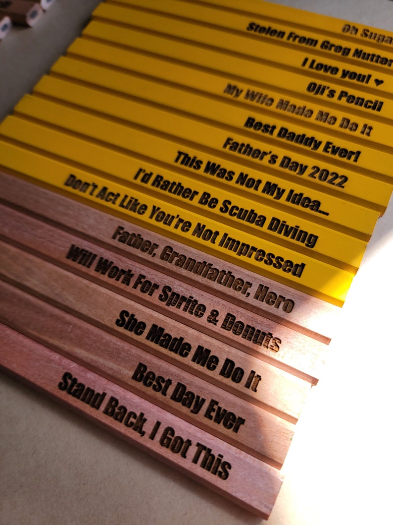 Puede incluir: Un conjunto de l&aacute;pices de madera amarillos y marrones con varios refranes humor&iacute;sticos grabados en ellos. Los refranes incluyen "Oh Suga", "Stolen From Greg Nutter", "I Love You!", "Oji's Pencil", "My Wife Made Me Do It", "Best Daddy Ever!", "Father's Day 2022", "This Was Not My Idea...", "Don't Act Like You're Not Impressed", "I'd Rather Be Scuba Diving", "Father, Grandfather, Hero", "Will Work For Sprite & Donuts", "She Made Me Do It", "Best Day Ever", y "Stand Back, I Got This".