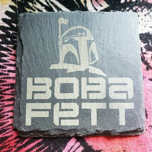 Pu&ograve; includere: Un sottobicchiere in ardesia nera con una silhouette bianca del casco di Boba Fett e il testo "BOBA FETT" in bianco.