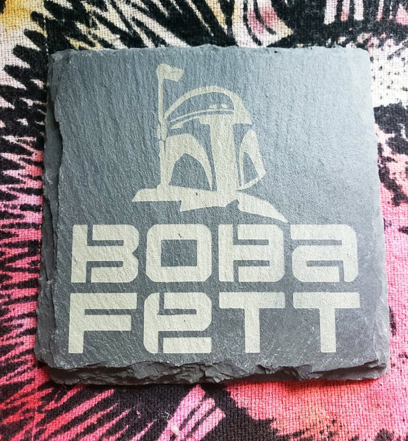 Pu&ograve; includere: Un sottobicchiere in ardesia nera con una silhouette bianca del casco di Boba Fett e il testo "BOBA FETT" in bianco.