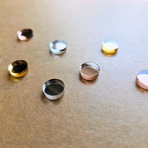 May include: Assortment of eight small round mirrored tiles in various colors, including gold, silver, rose gold, and black. The tiles are arranged on a brown surface.