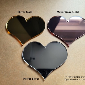 May include: Three heart-shaped acrylic mirrors in gold, rose gold, and silver. The mirrors are one-sided, with a solid gray back.