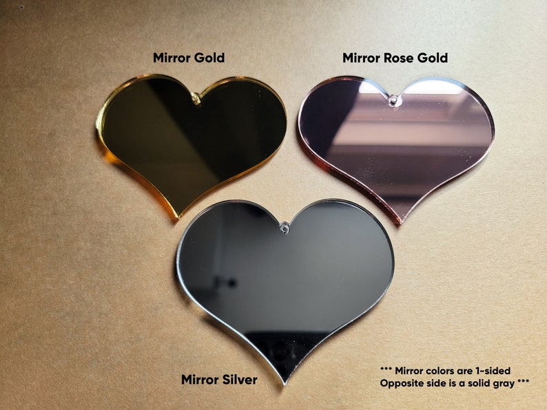 May include: Three heart-shaped acrylic mirrors in gold, rose gold, and silver. The mirrors are one-sided, with a solid gray back.
