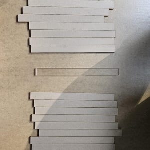 May include: Arrangement of rectangular, light gray cardboard pieces. Two stacks of the cardboard pieces are visible, with a single, clear, rectangular piece between them. The pieces are arranged on a light brown surface.