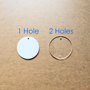 May include: Two round acrylic blanks, one white and one clear. The white blank has one hole, and the clear blank has two holes. The text "1 Hole" and "2 Holes" is printed above the blanks.