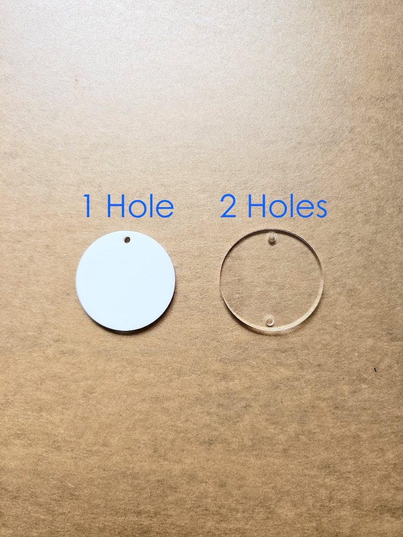 May include: Two round acrylic blanks, one white and one clear. The white blank has one hole, and the clear blank has two holes. The text "1 Hole" and "2 Holes" is printed above the blanks.