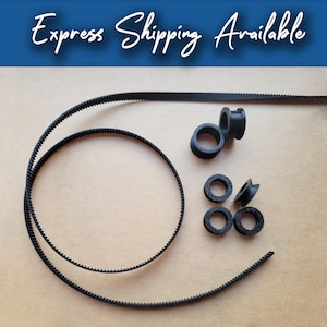 以下が含まれることがあります： 黒いタイミングベルトとプーリーが薄茶色の表面に配置されています。ベルトは歯付きのデザインで柔軟性があります。「Express Shipping Available」の文字が画像上部にあります。