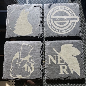 May include: Four black slate coasters with white etched designs. The designs include a wolf, a smiling face with the text "I was one of those deaf-mutes", a creature with a red box, and a leaf with the text "NERV".