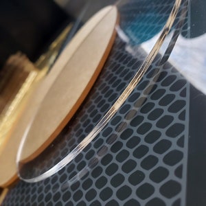May include: A clear acrylic round coaster with a wood veneer edge. The coaster is on a black surface with a textured pattern.