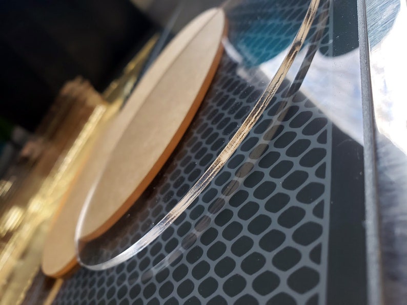 May include: A clear acrylic round coaster with a wood veneer edge. The coaster is on a black surface with a textured pattern.