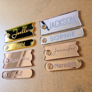 May include: A set of seven personalized acrylic name tags with rounded corners. The tags are in gold, silver, and rose gold colors with names written in a cursive font. The names are "Joelle", "Tracey", "Khavina", "Chanell", "Jackson", "Sophie", and "Jennifer".