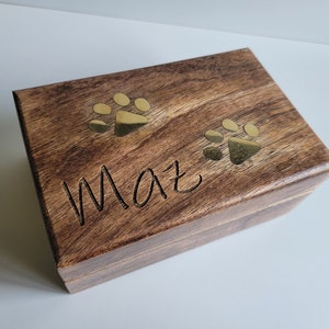 Puede incluir: Una caja de madera con tapa con bisagras, grabada con el nombre "Max" y dos huellas de patas doradas.