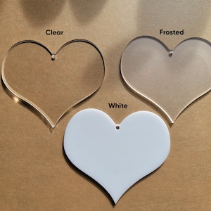 May include: Three heart-shaped acrylic blanks in different finishes: clear, frosted, and white. Each blank has a hole at the top for hanging.