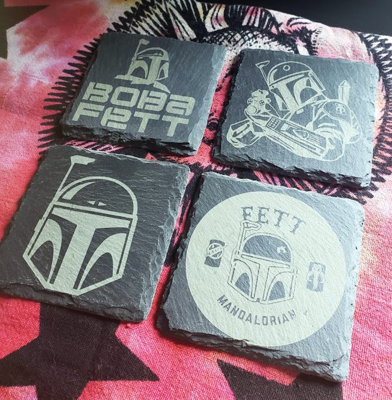 Pu&ograve; includere: Quattro sottobicchieri in ardesia nera con un design bianco che presenta il personaggio di Star Wars Boba Fett. I sottobicchieri presentano diversi design, tra cui un ritratto di Boba Fett, le parole "Boba Fett" e un design circolare con le parole "Fett Mandalorian".