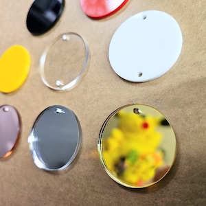 May include: Assortment of round acrylic blanks in various colors and finishes, including clear, gold, silver, black, white, and pink. Each blank has a hole for hanging or attaching.