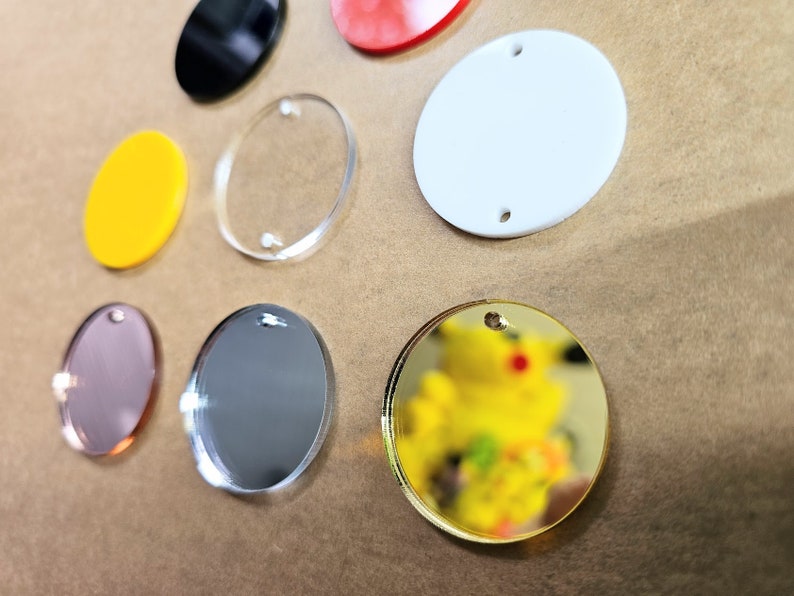 May include: Assortment of round acrylic blanks in various colors and finishes, including clear, gold, silver, black, white, and pink. Each blank has a hole for hanging or attaching.