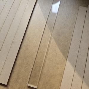 May include: A transparent, rectangular acrylic strip lies diagonally, with several light brown, rectangular wooden strips around it. The acrylic reflects light, contrasting with the matte wood. The items are arranged on a neutral-toned surface.